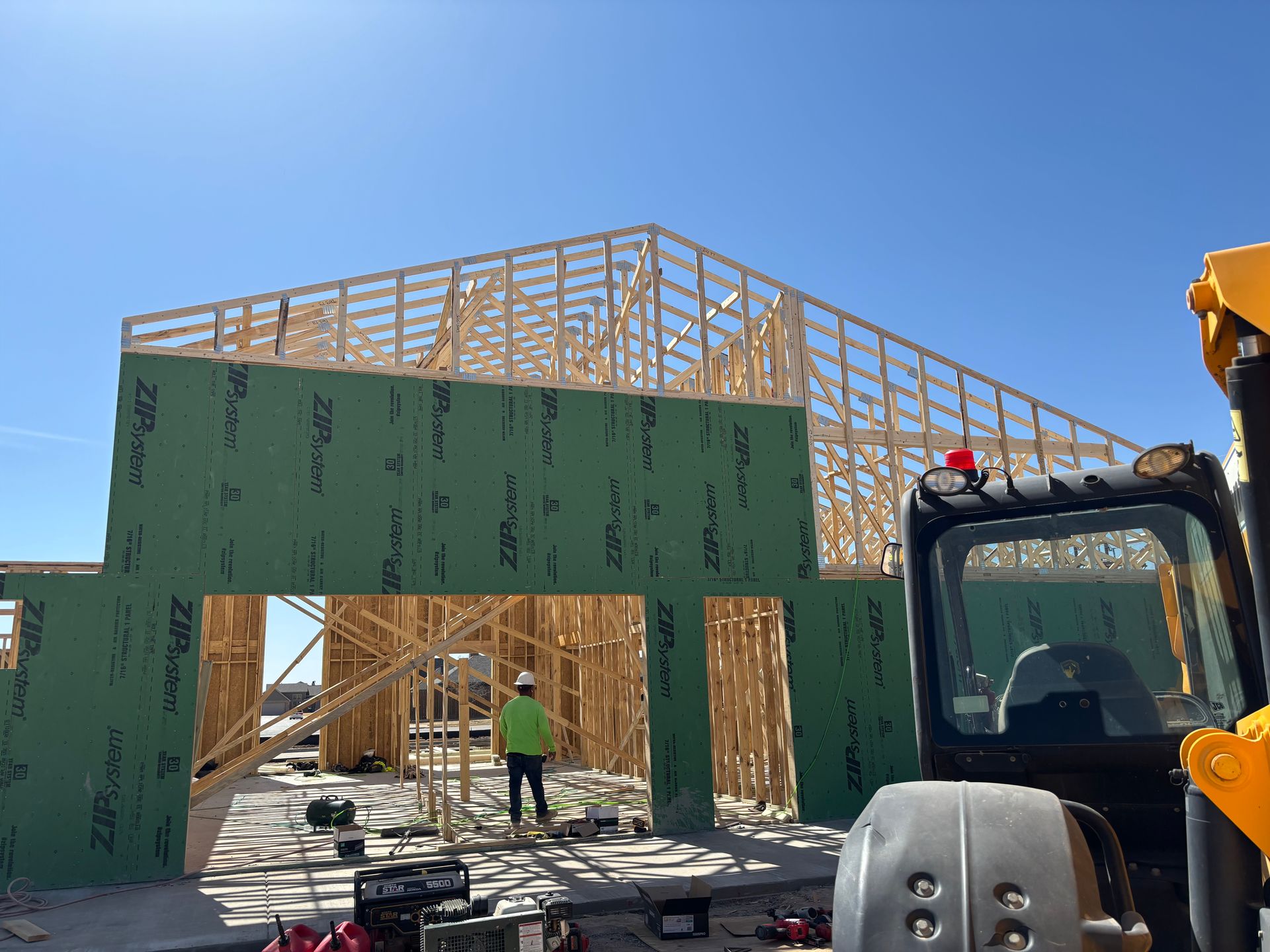 House under construction with wooden roof framing, green sheathing, and workers on site under a blue sky