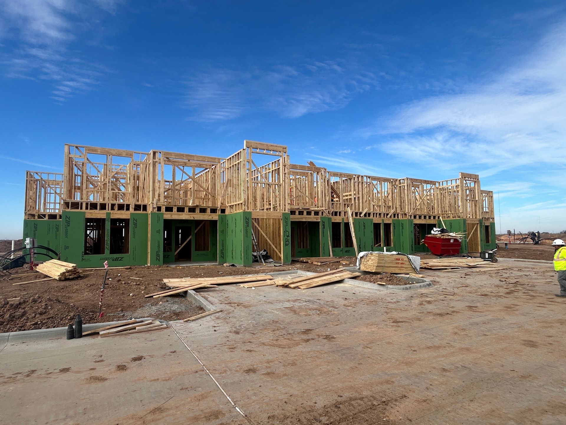 Partially built wooden house frame on a dirt lot under a blue sky, with construction materials scattered nearby