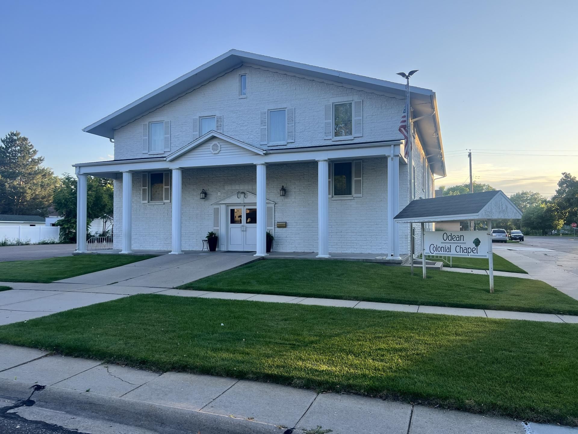 Two-story white building with columns, a sign in the grass, and a paved walkway.