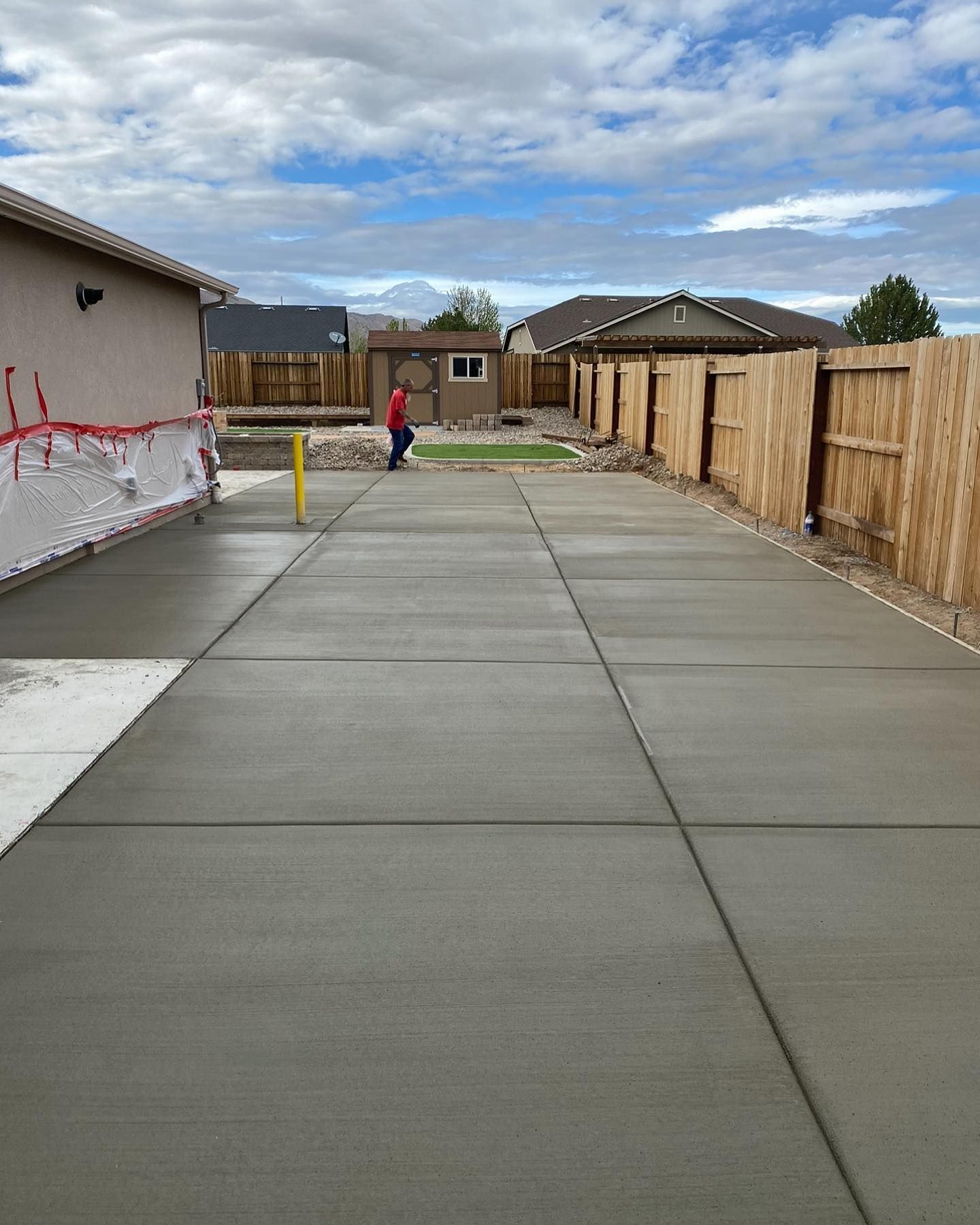 A person stands at the end of a long, newly poured concrete walkway in a residential backyard with a wood fence.
