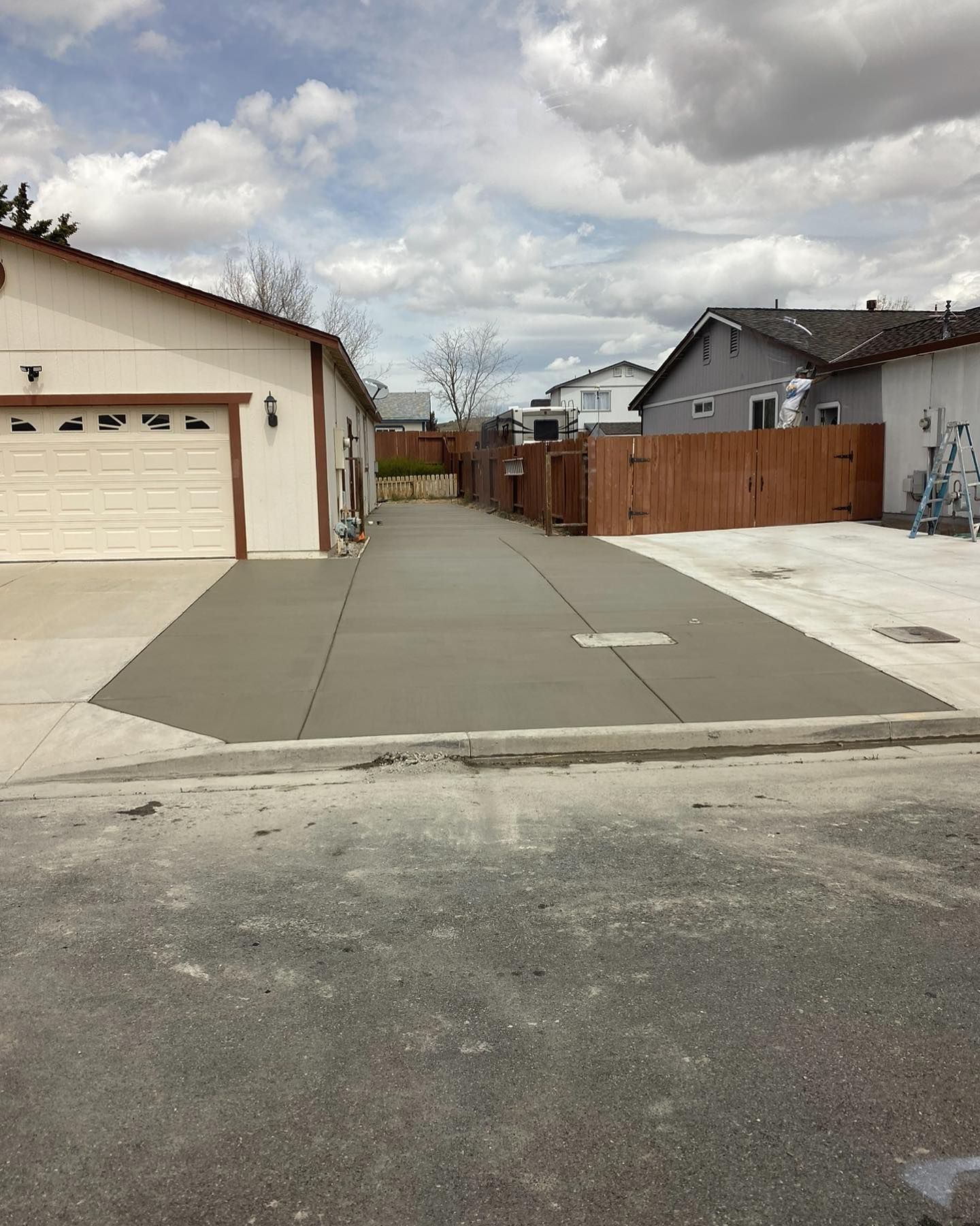 A newly poured concrete driveway leads from a suburban street toward a garage and a wooden fence between two houses.