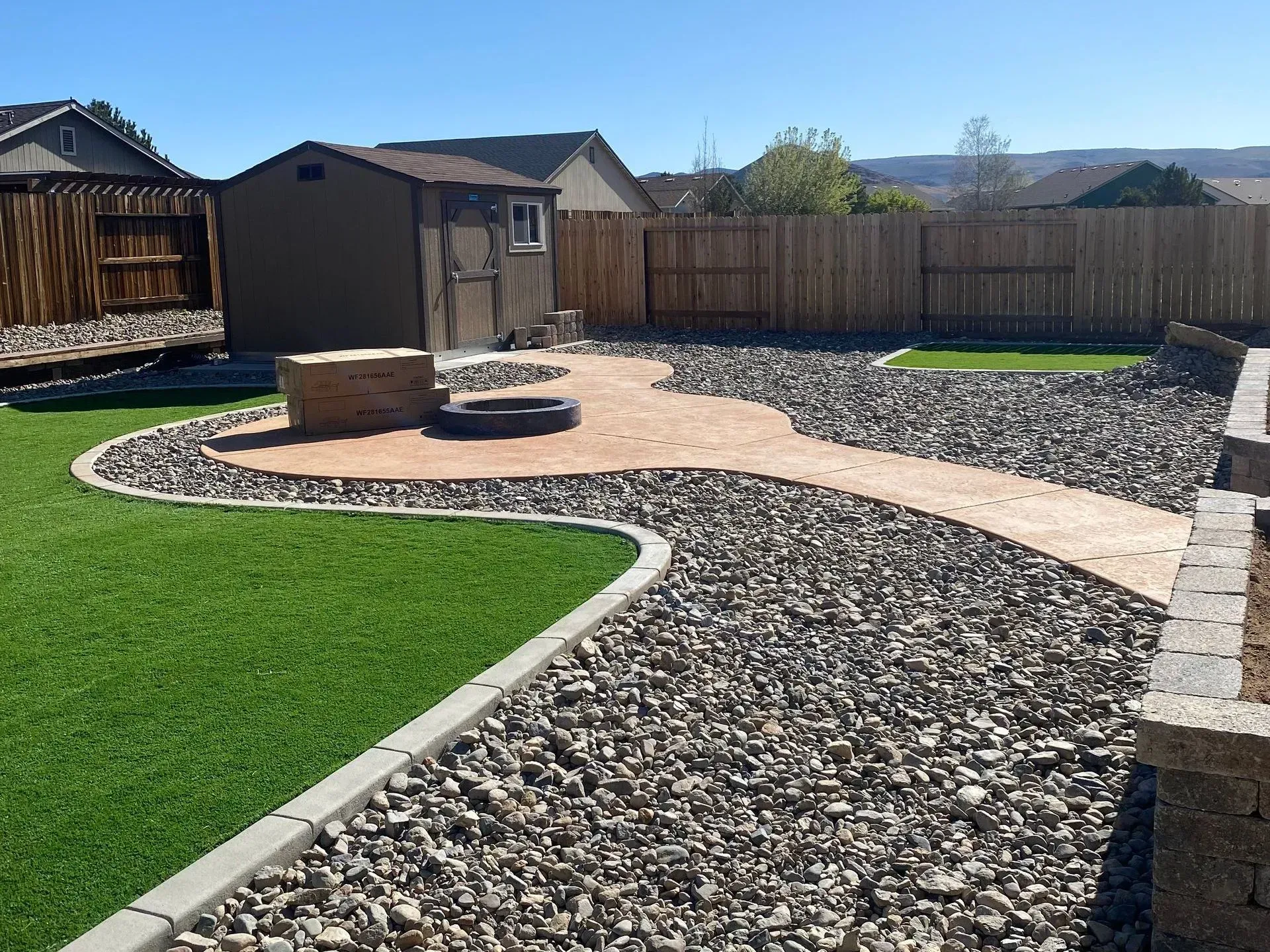 A backyard with a green artificial turf lawn, a gravel border, and a paved patio area featuring a fire pit and a shed.
