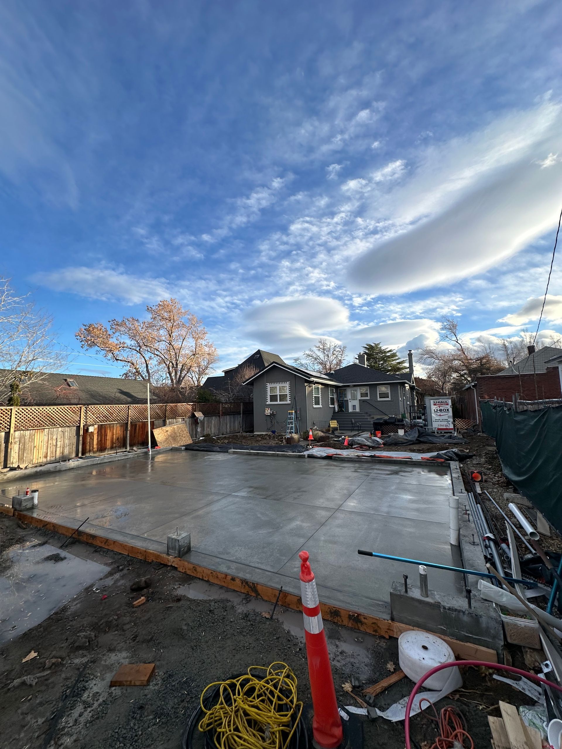 A freshly poured concrete foundation for a building project under a bright, cloudy sky, with construction debris nearby.