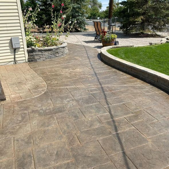 A stamped concrete patio featuring tan rectangular pavers, curved stone walls, and a manicured green lawn in a backyard.