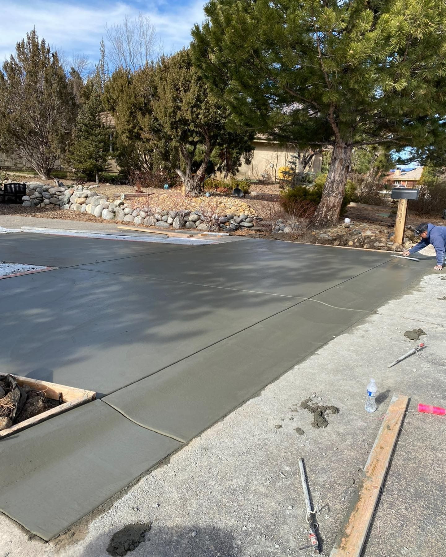 A worker smooths a newly poured concrete driveway outdoors in a sunny, residential setting with trees in the background.