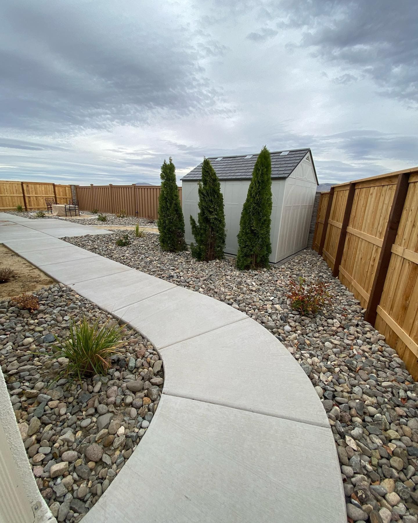 A curved concrete walkway leads through a landscaped yard filled with decorative rocks toward a shed and wooden fences.