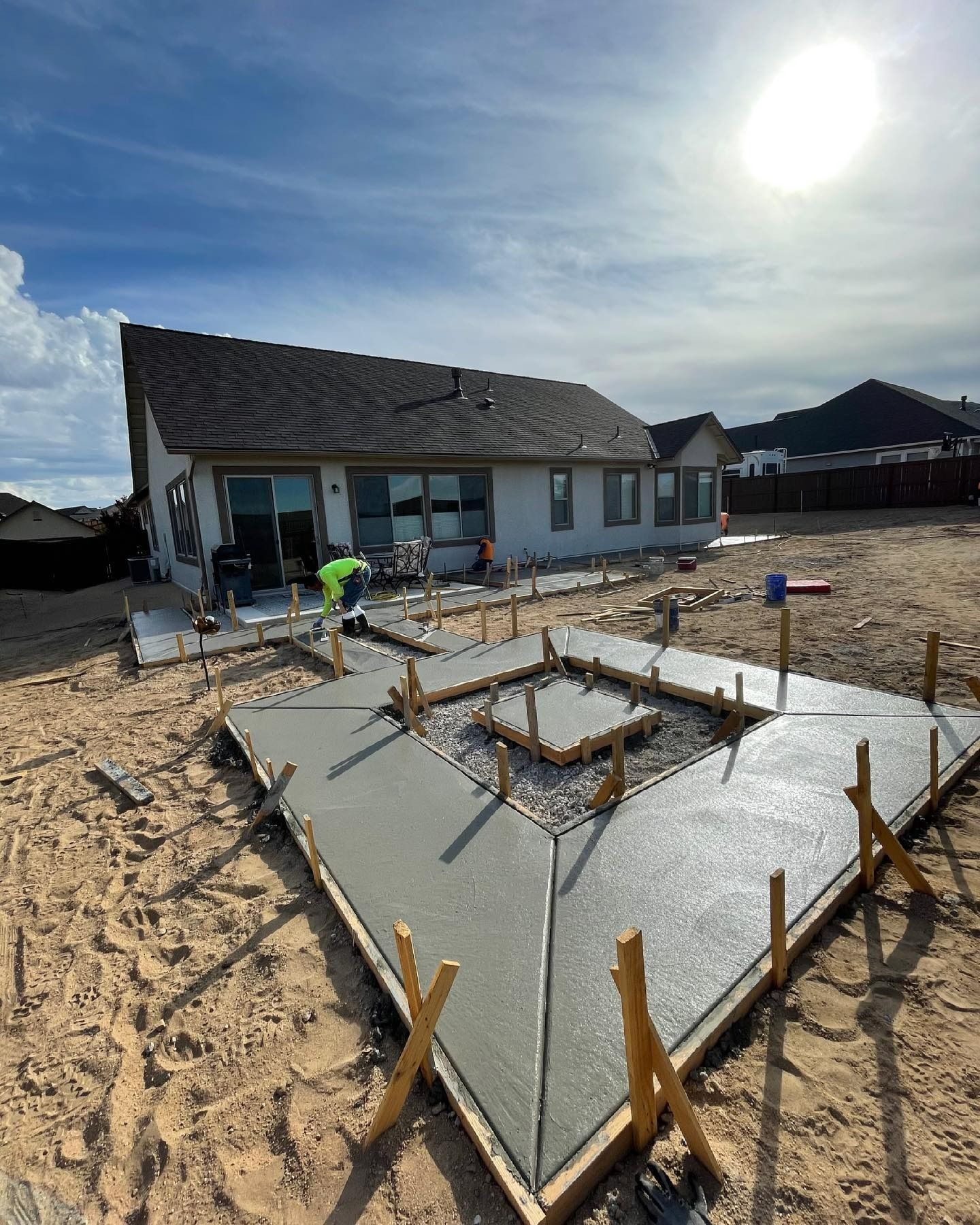 A worker finishes a wet concrete patio with a central square fire pit frame in a backyard under a sunny, blue sky.