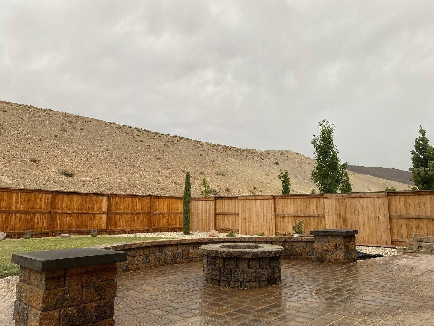 A stone fire pit and seating wall on a paved patio, set against a wooden fence and a large, dry desert hill.