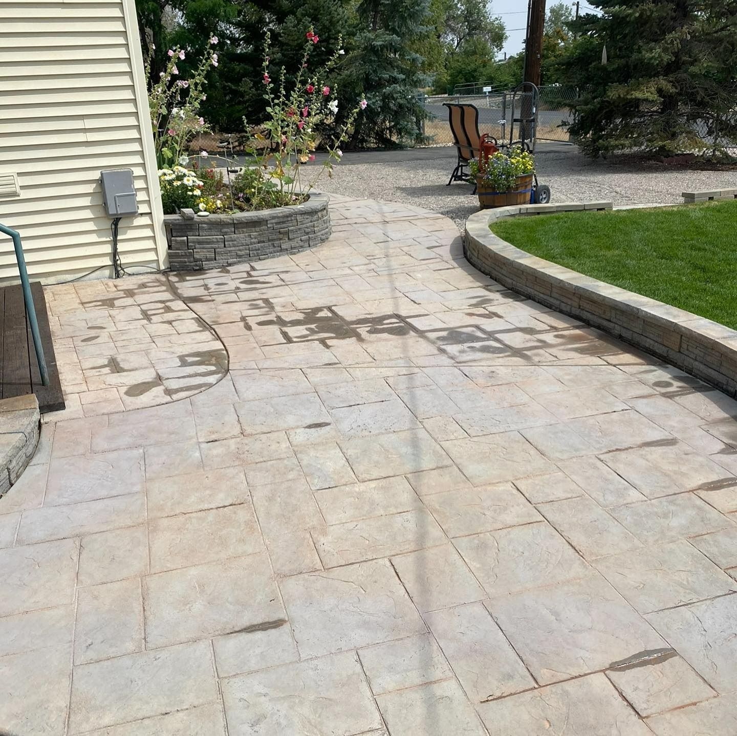 A beige paver patio with a curved stone retaining wall bordering a grassy area next to a light-sided house.