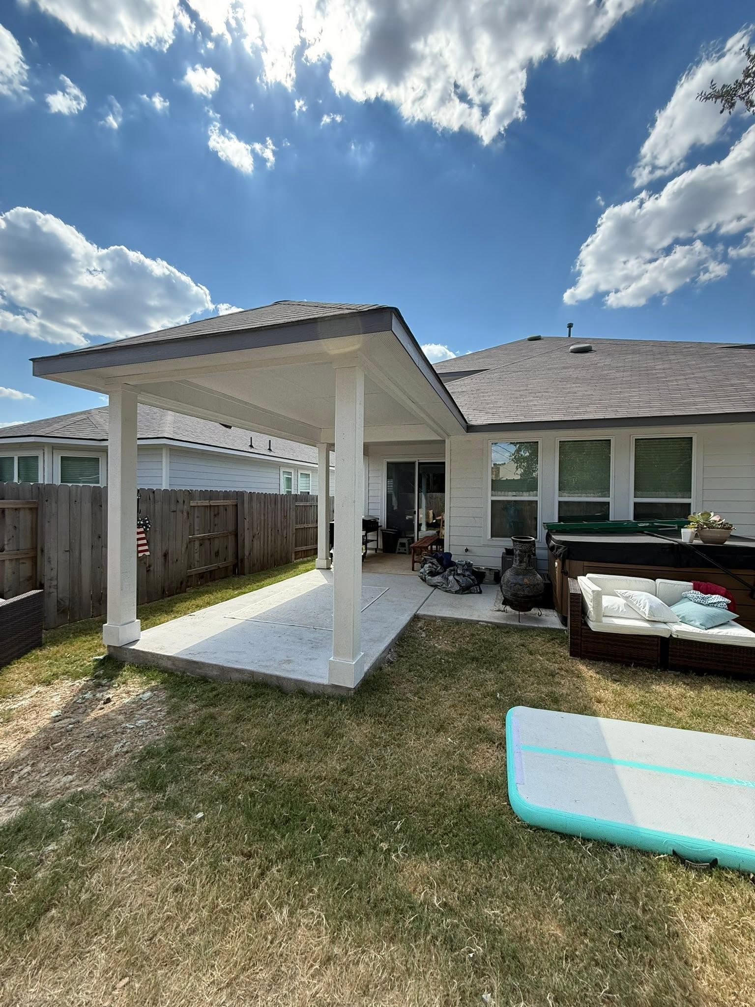 Backyard patio with white pergola, lawn, fence, and a house under a partly cloudy sky