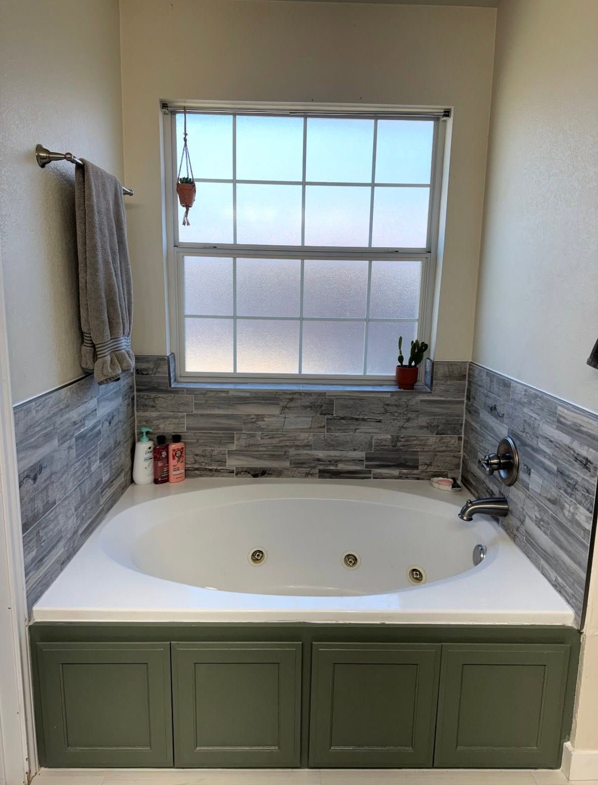 Bathroom with white corner tub, green cabinets, gray tile walls, and frosted window above.