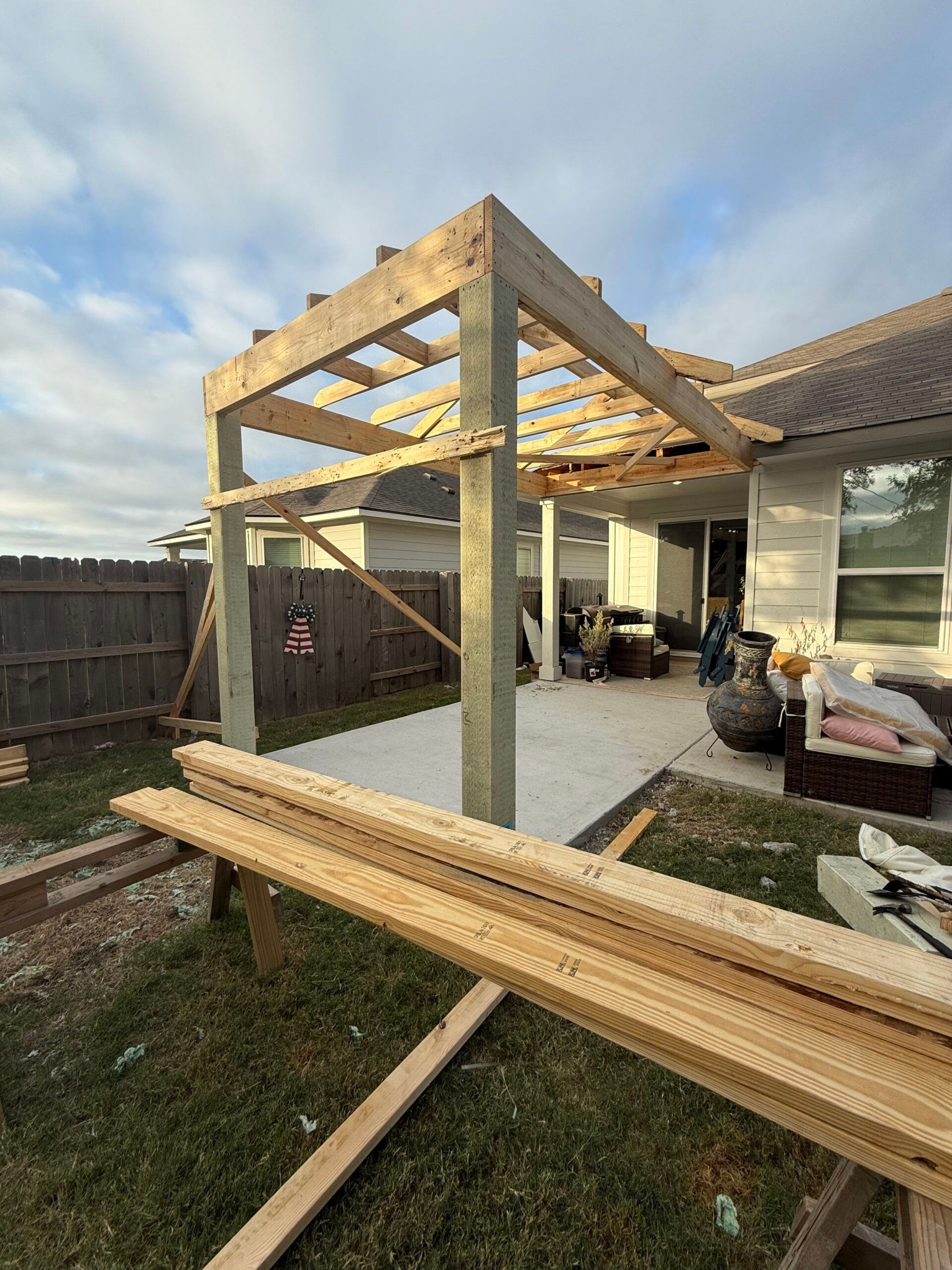 Wooden pergola frame under construction beside a house with lumber stacked in the yard