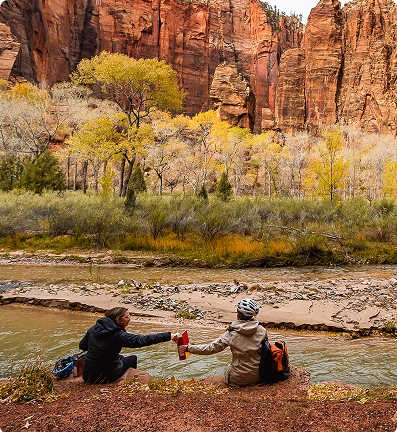 Two people seated by a river in Zion National Park, sharing a drink; red rock canyon backdrop.