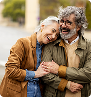 Smiling couple embracing outdoors. Woman leans on man's shoulder. Both are wearing jackets.