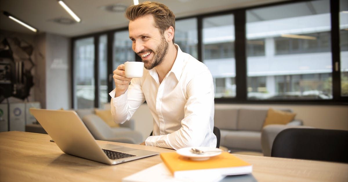 Um homem está sentado a uma mesa tomando uma xícara de café enquanto usa um laptop.