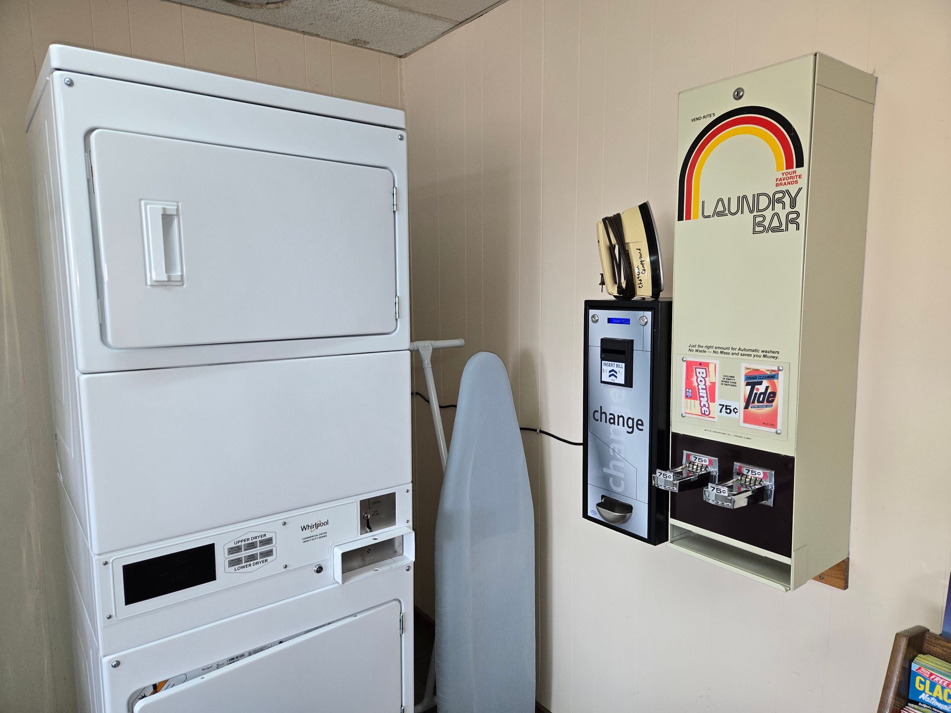 Stacked white washer and dryer, ironing board, and laundry coin machine.