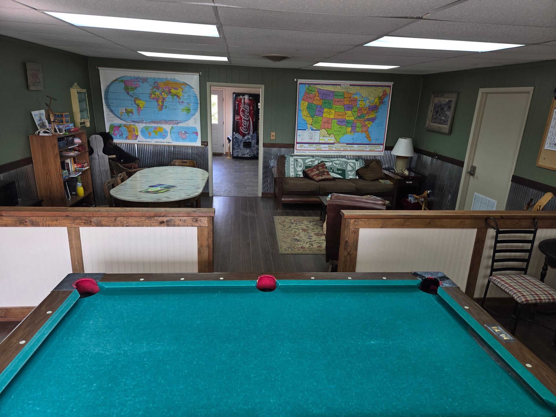 A game room with a pool table in the foreground, maps on the walls, and a sitting area beyond.