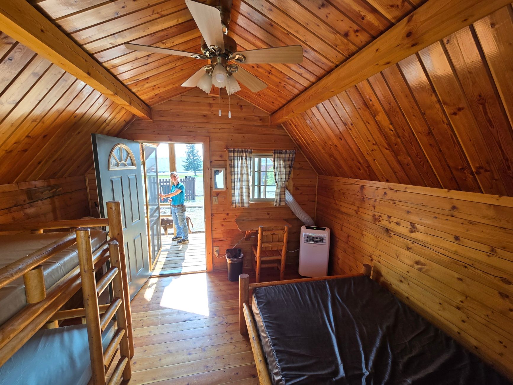 Interior of a wood cabin with bunk beds, a single bed, open door, and a person standing outside.