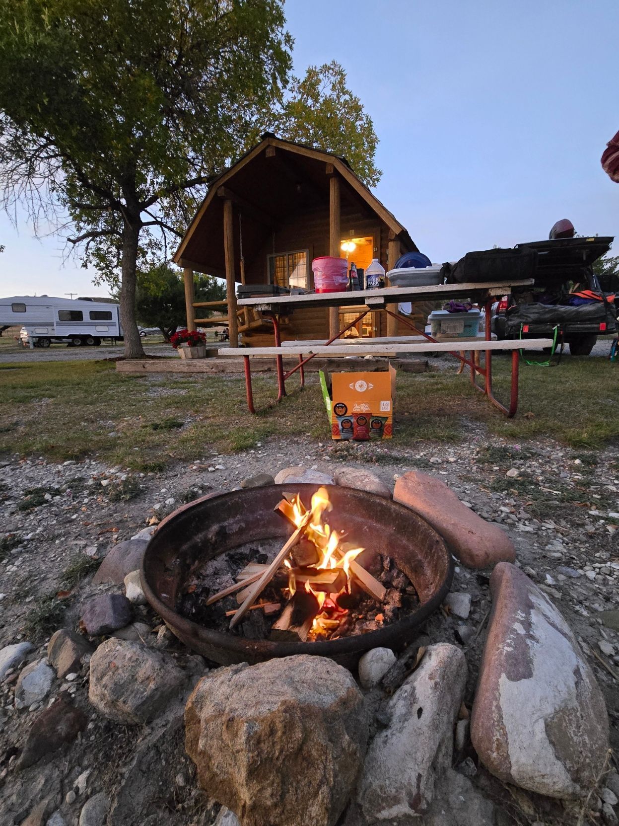 Campfire burning, cabin and picnic table in the background. Camping scene at dusk with an RV parked nearby.