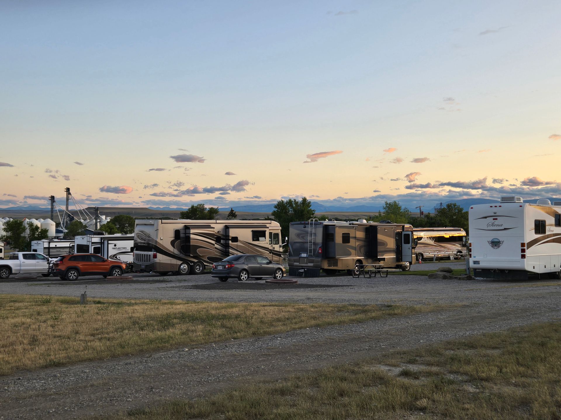 RVs and cars parked in a campground at dusk, with a soft orange and blue sky and mountains in the distance.