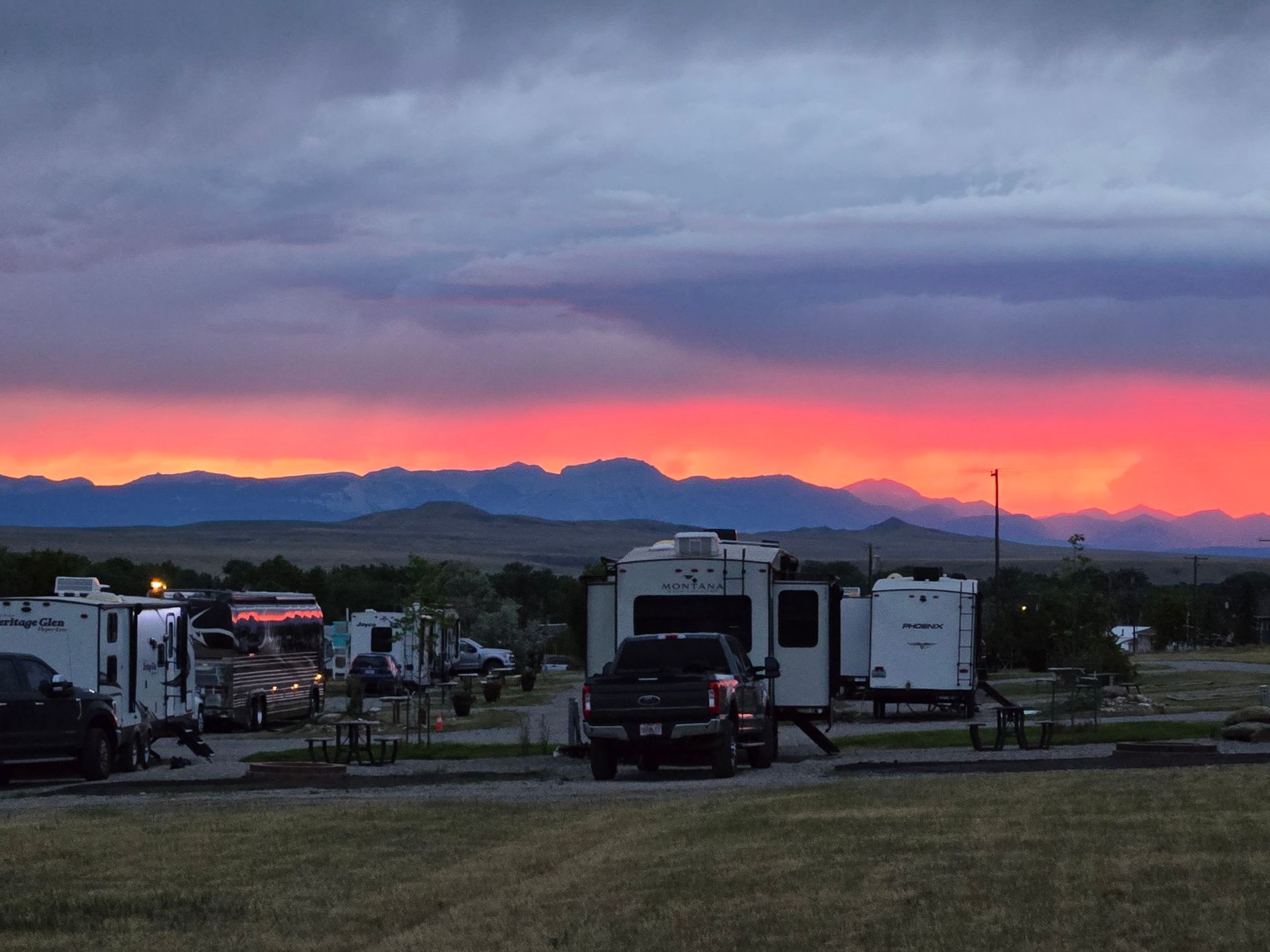 Campground at sunset, RVs and trucks parked, with colorful sky over distant mountains.