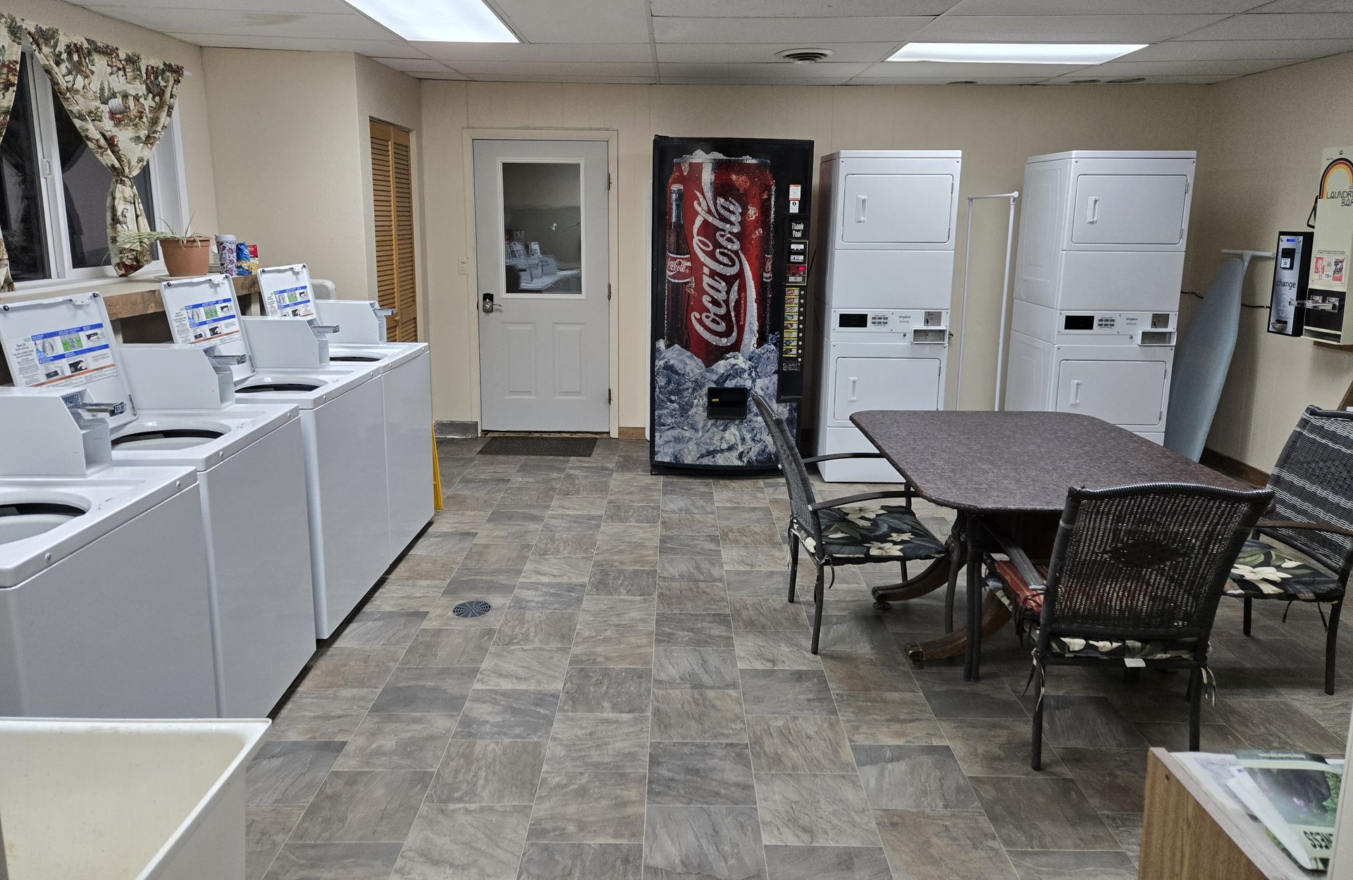 A laundromat with washing machines, dryers, a vending machine, a table with chairs, and a door.