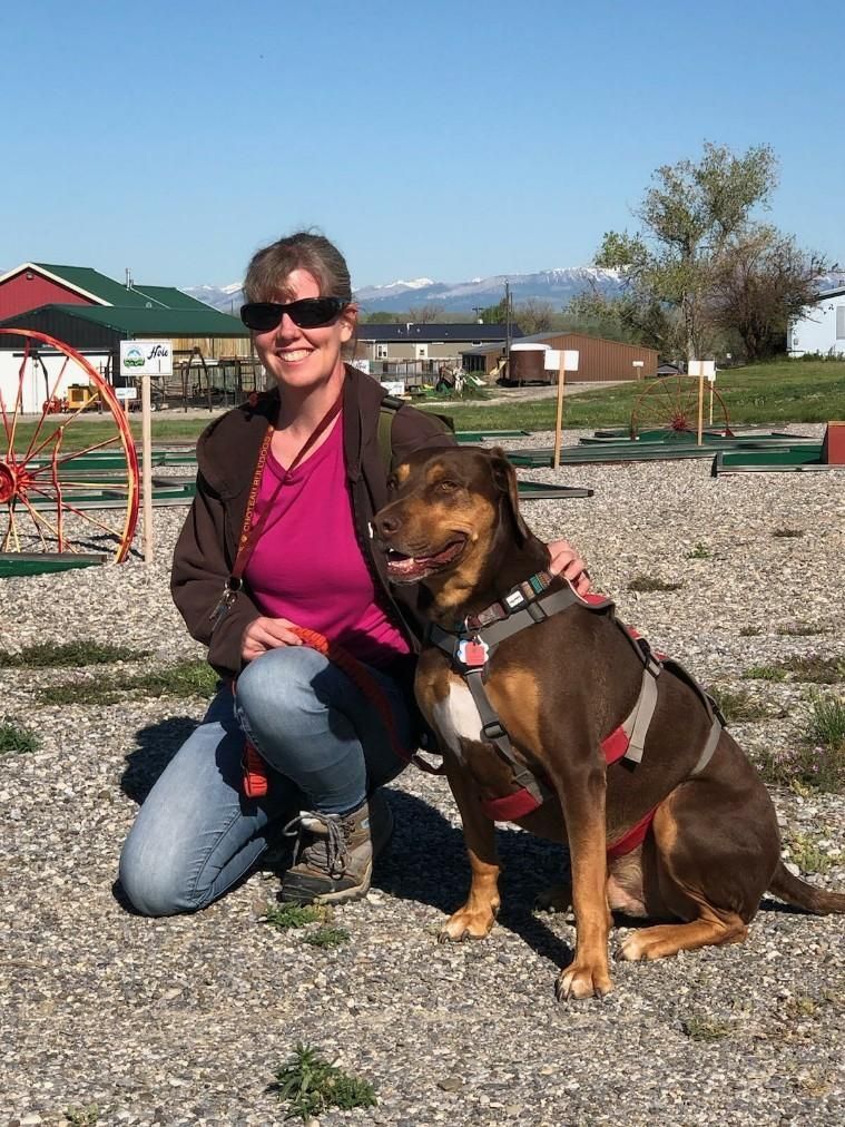 Woman kneels beside a brown dog wearing a harness outdoors. They are smiling.