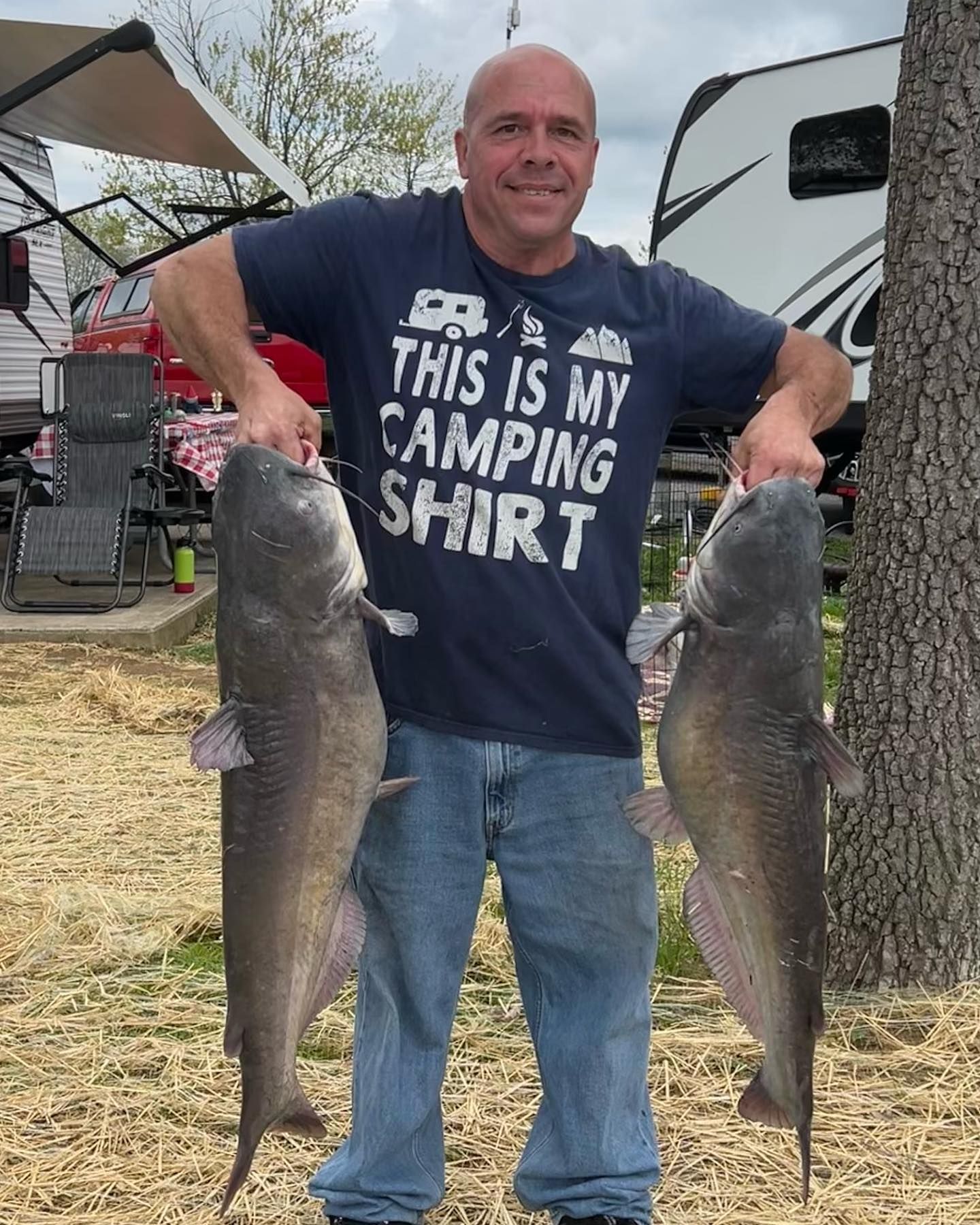 Smiling man outdoors holding two large fish, wearing a navy camping shirt and jeans.