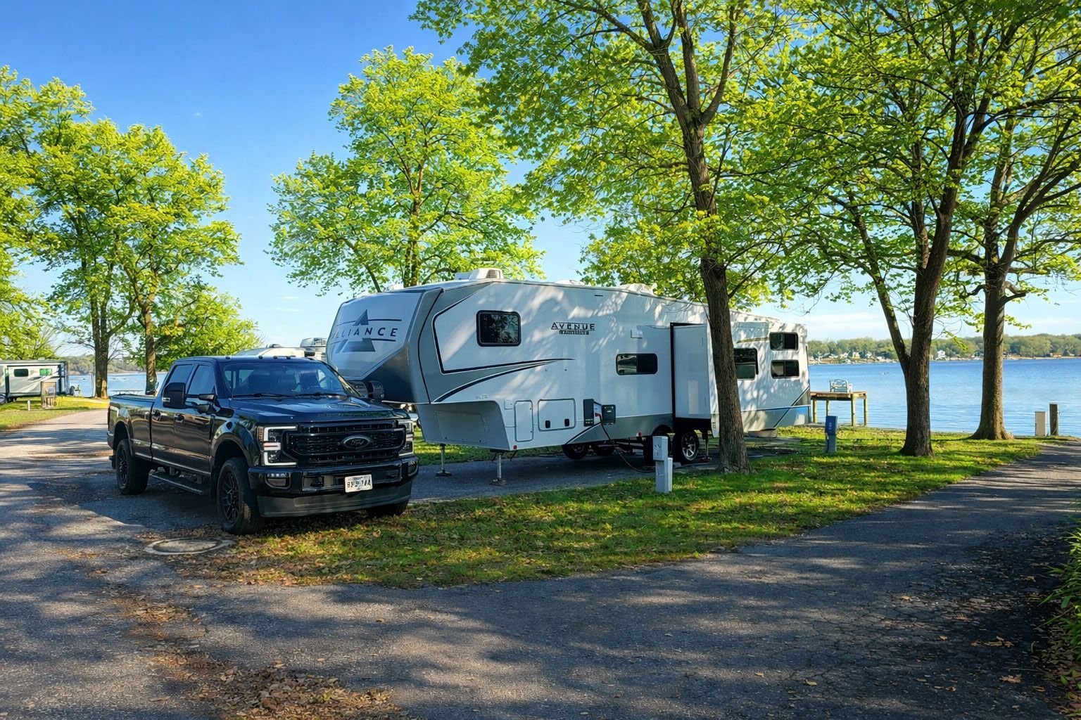 Black pickup and white camper parked by a lakeside under green trees on a sunny day