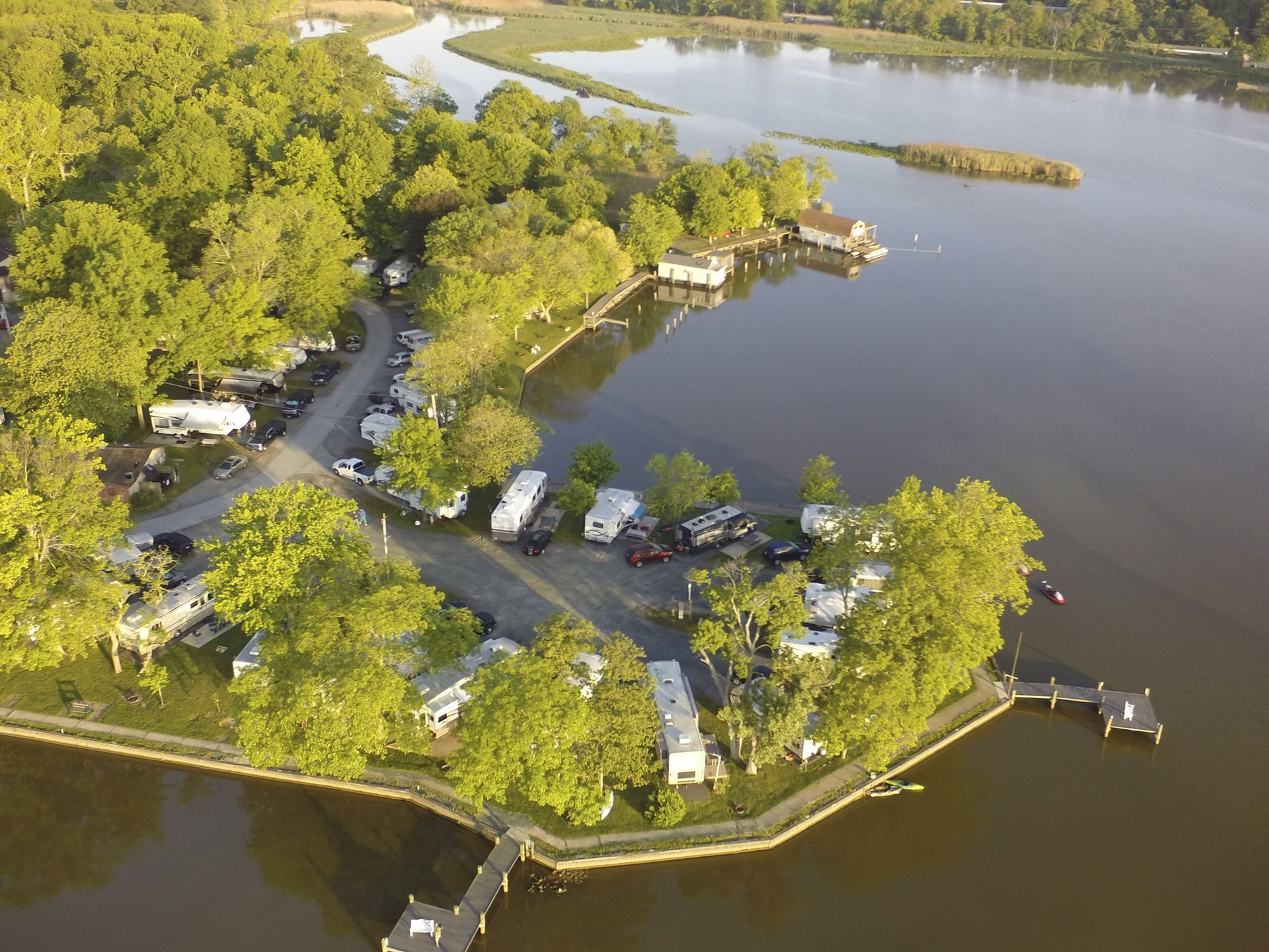Aerial view of a tree-lined lakeside park with a winding road, dock, and small buildings on the shore.