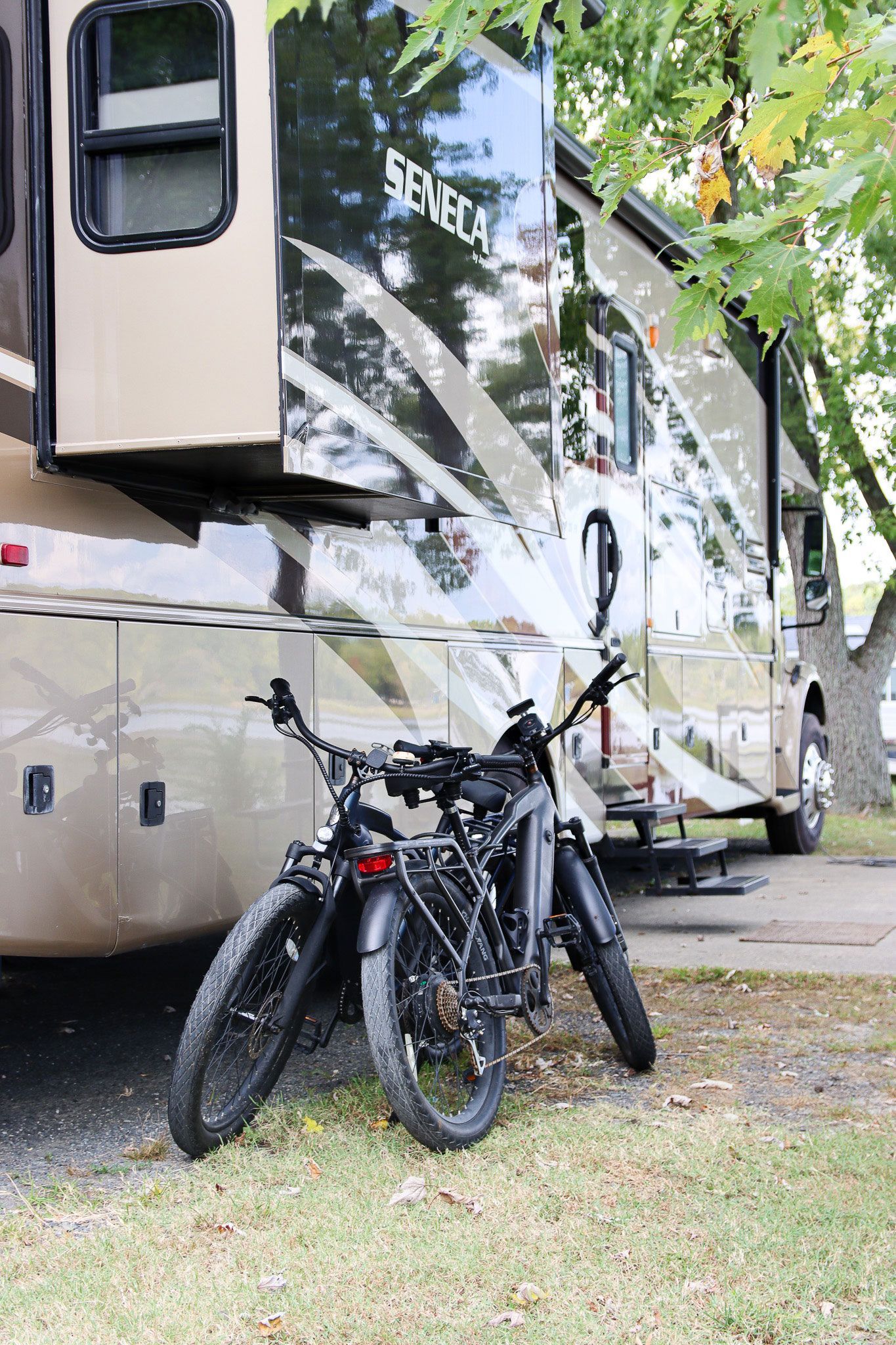 Two bicycles parked beside a beige RV under leafy trees.
