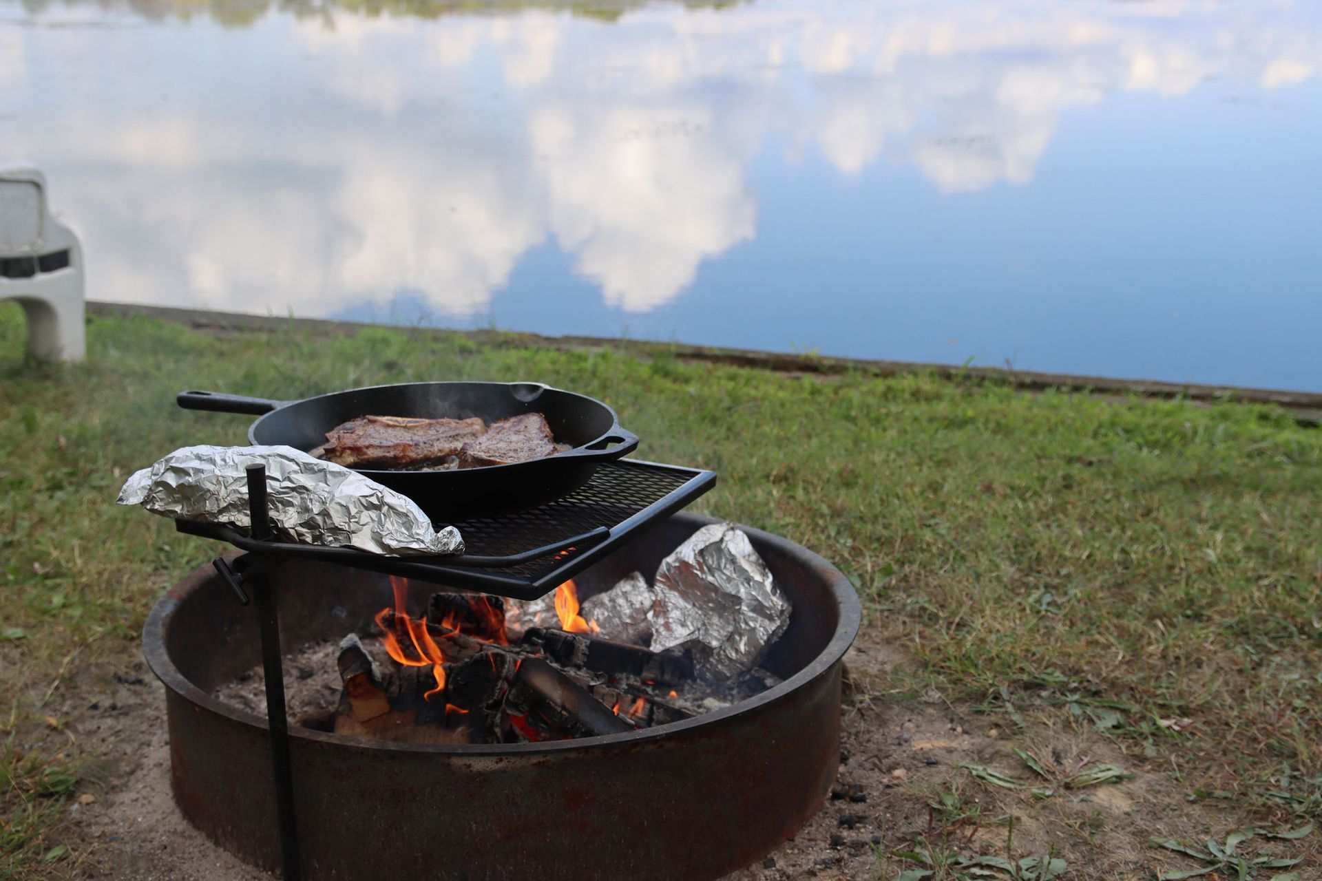 A campfire in a metal ring with a black grill pan cooking meat by a lakeside