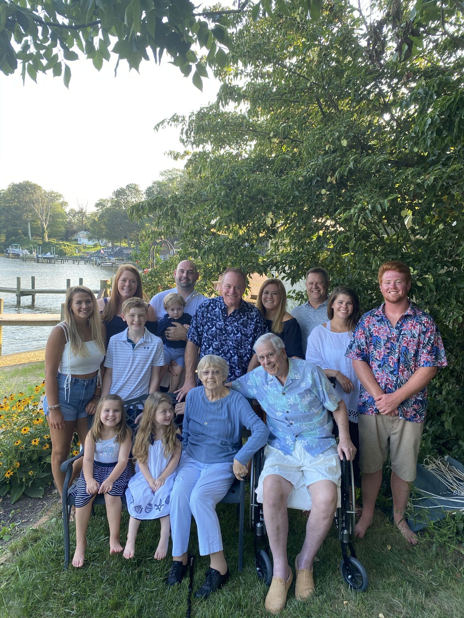 Group of people posing outdoors under trees, smiling beside a lakeside fence at sunset.