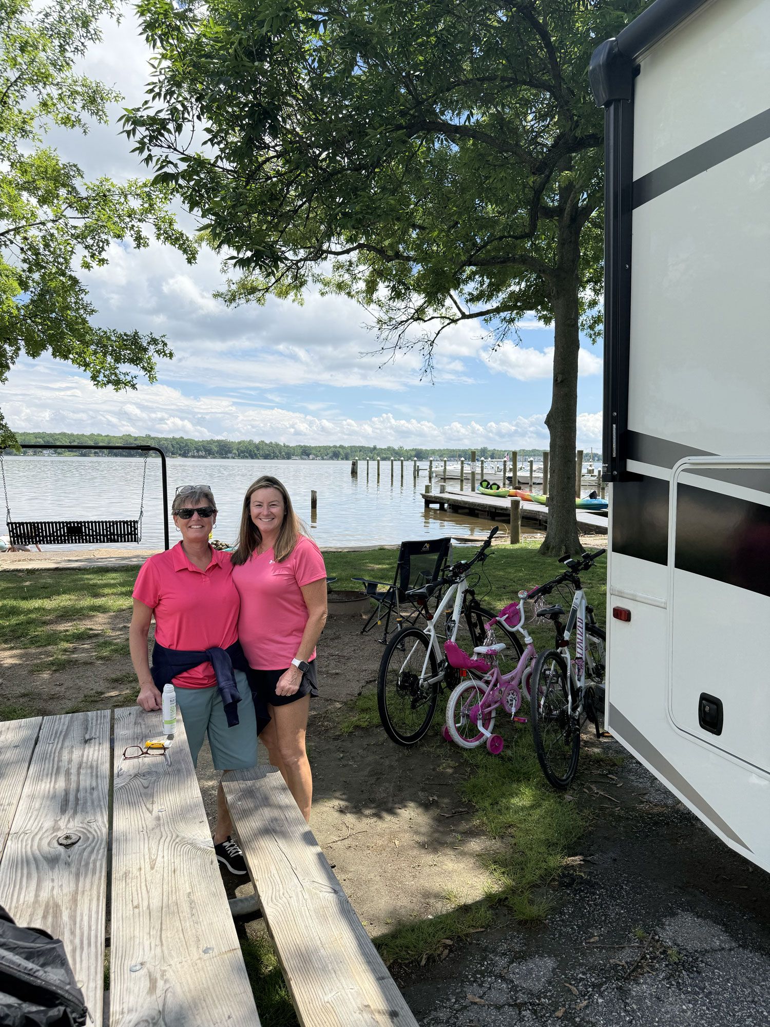 Two people stand on a dock by a lake with bikes, trees, and an RV nearby.
