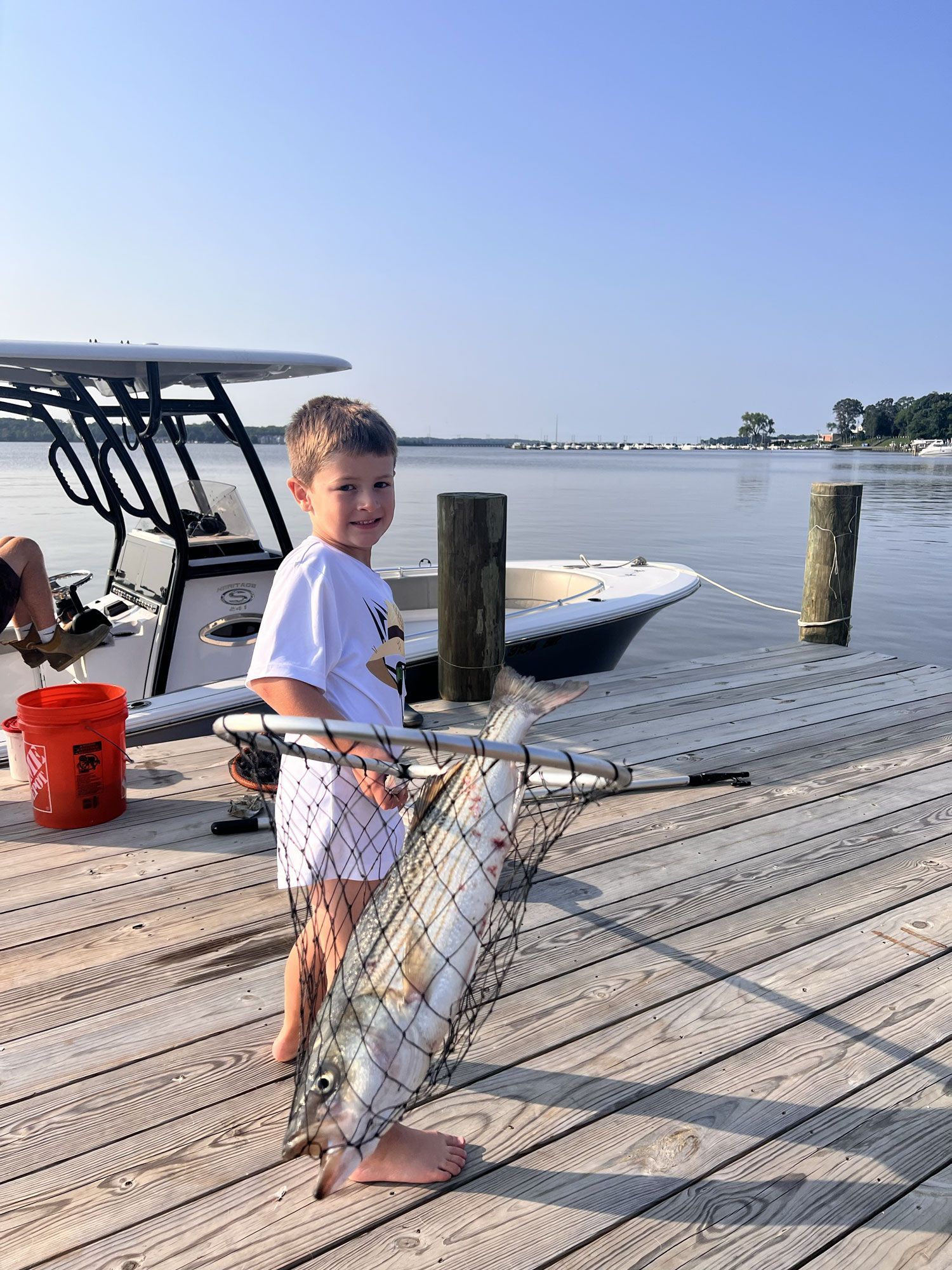 Boy holding a fish on a dock by the water, with a boat and fishing gear nearby.