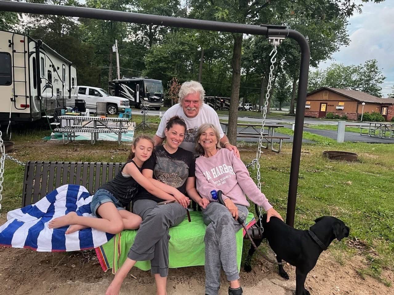 Three people sit on a green bench under a frame, smiling; a black dog stands nearby outdoors.
