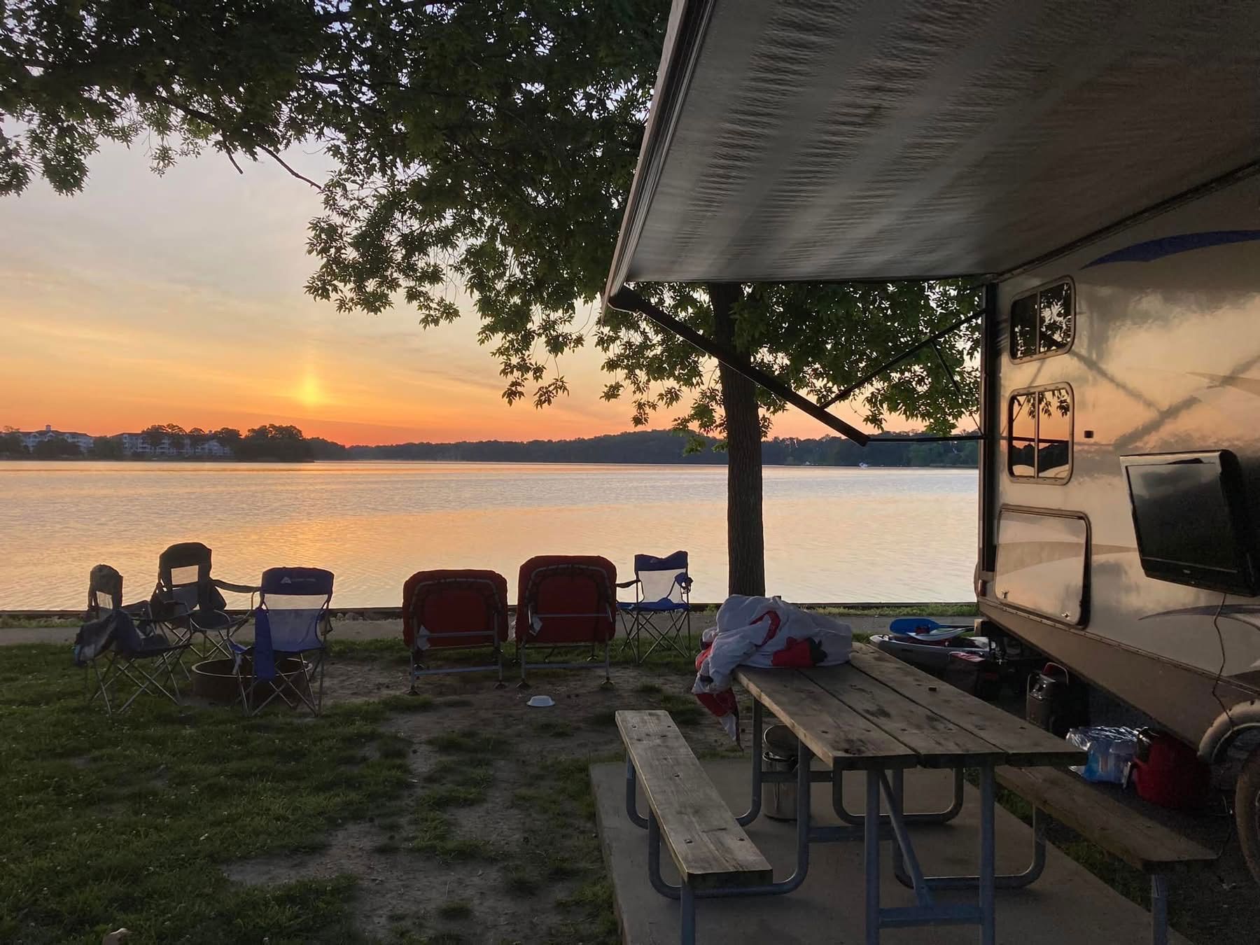 Sunset over a lake with chairs, picnic tables, and a camper at a campsite