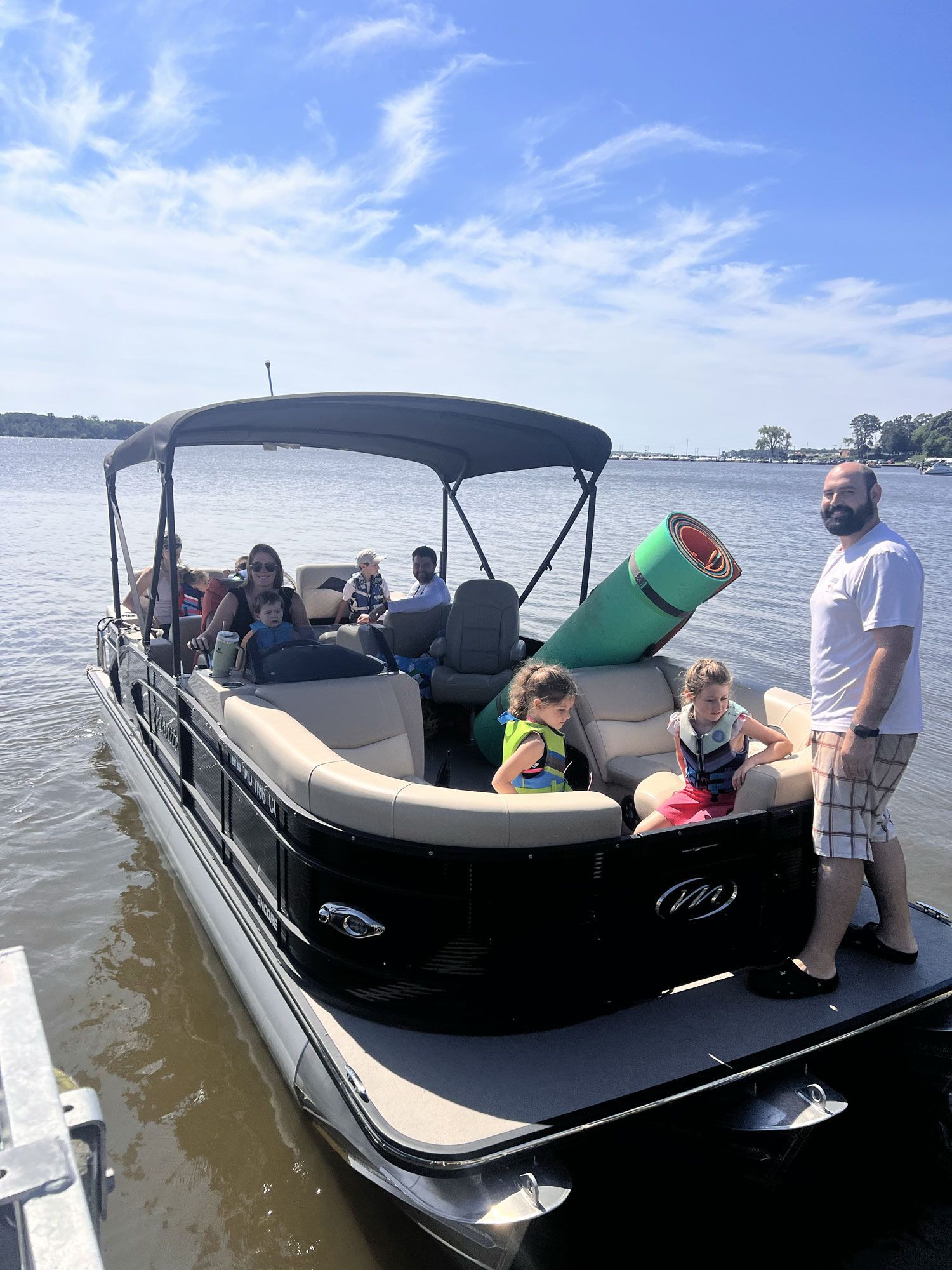 People on a black pontoon boat on a lake, with a bright green float and blue sky overhead.