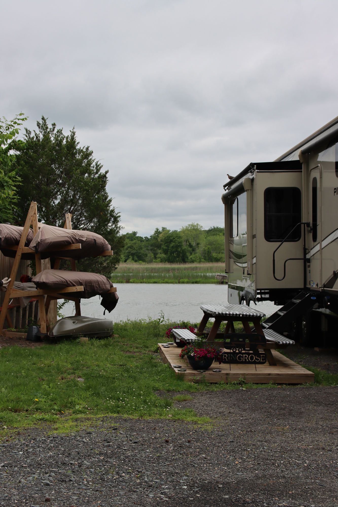 RV campsite by a lake with picnic table, grill, flower pot, and stacked canoes on the left