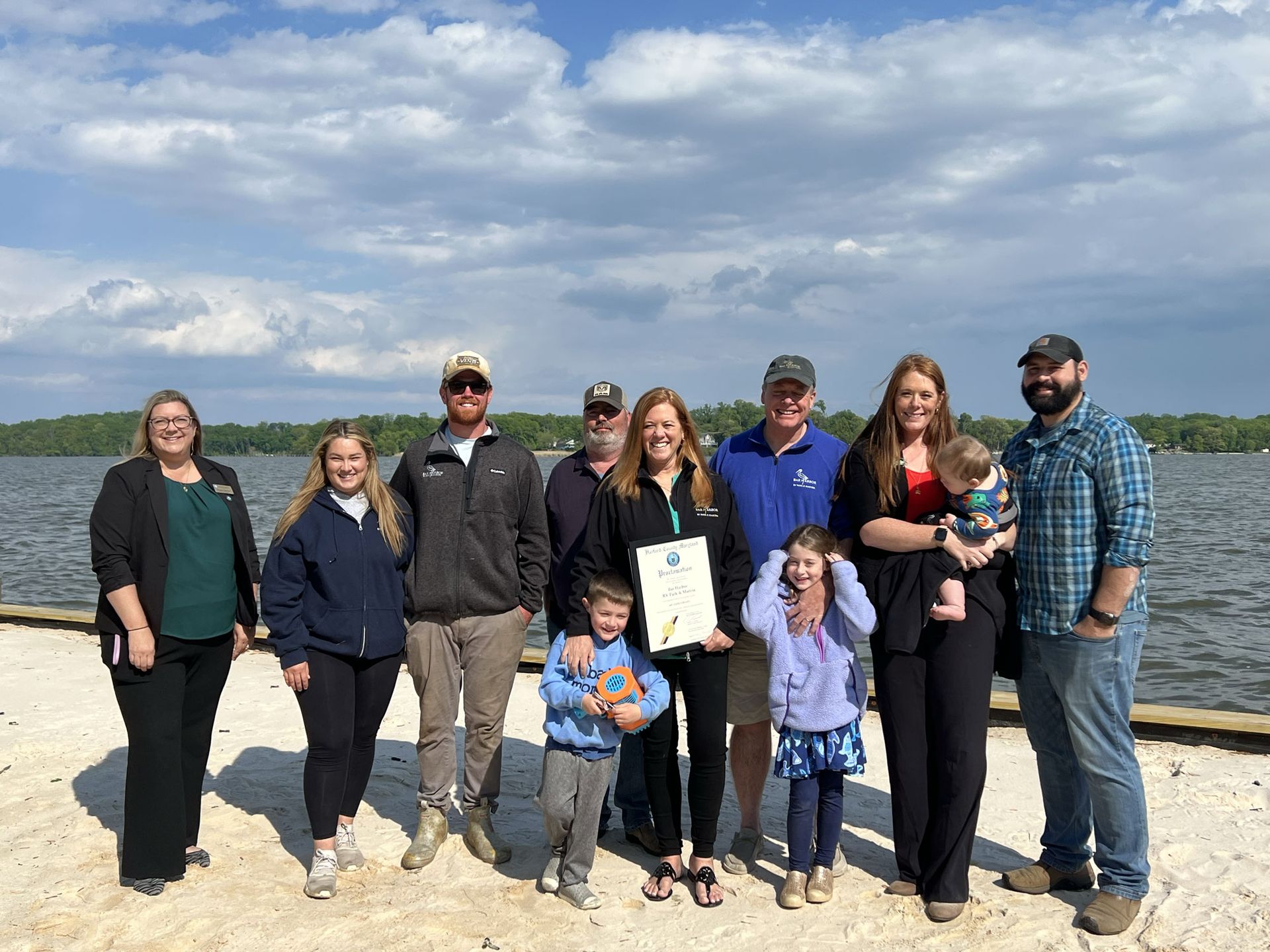 Group of people posing on a sandy beach, holding a certificate, with water and cloudy sky behind them.