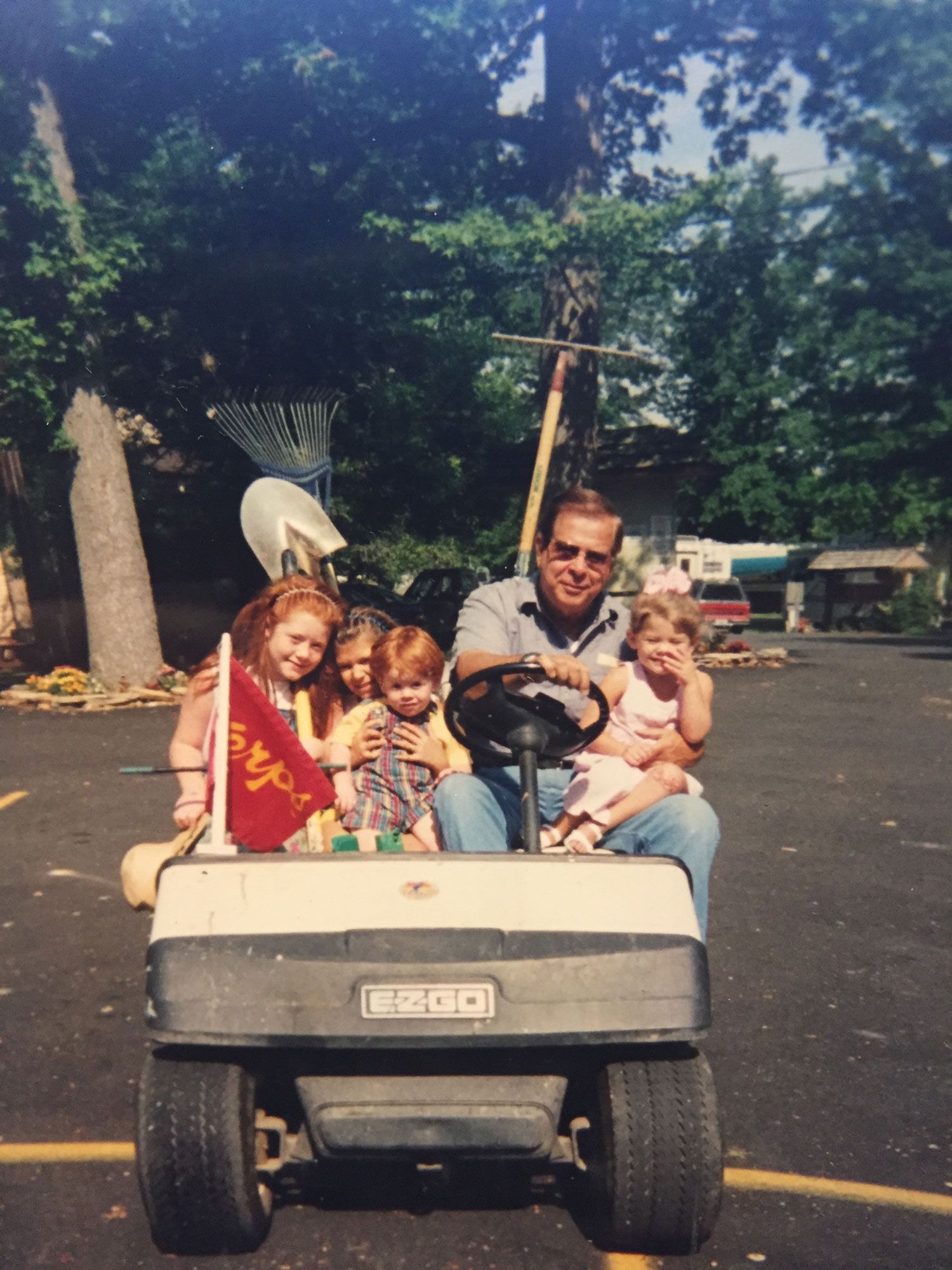 Family riding together on a golf cart outdoors in a parking lot, with trees in the background