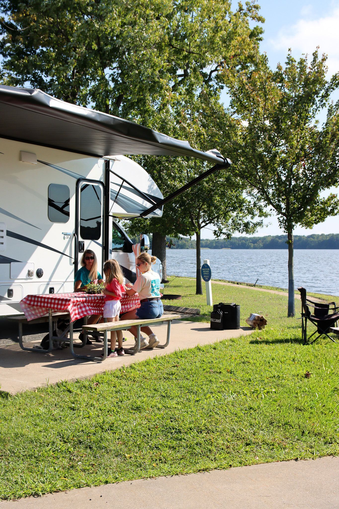 People sitting at a picnic table beside an RV by a lakeshore on a sunny day