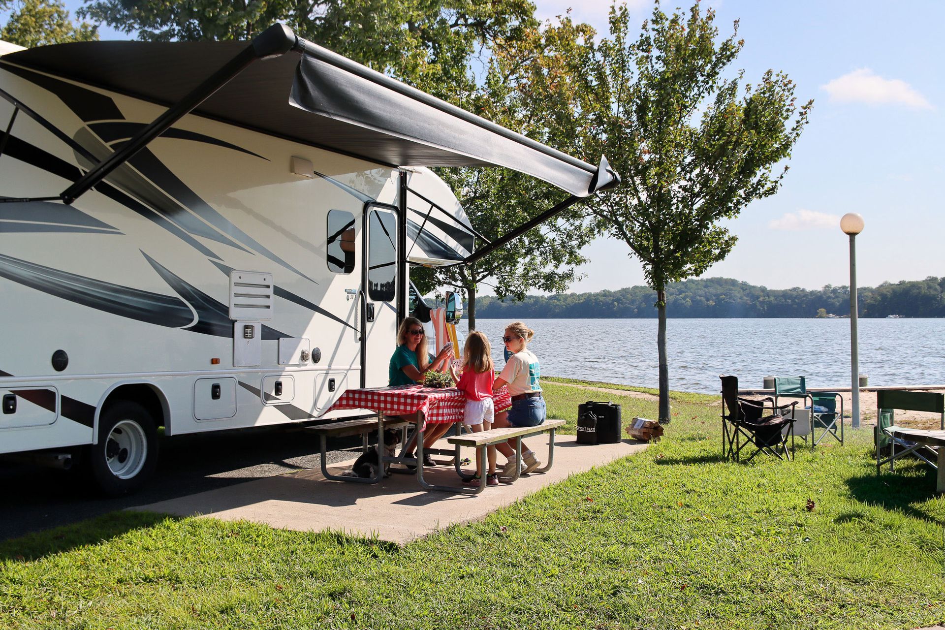 RV campsite by a lake with people relaxing at a picnic table under an awning