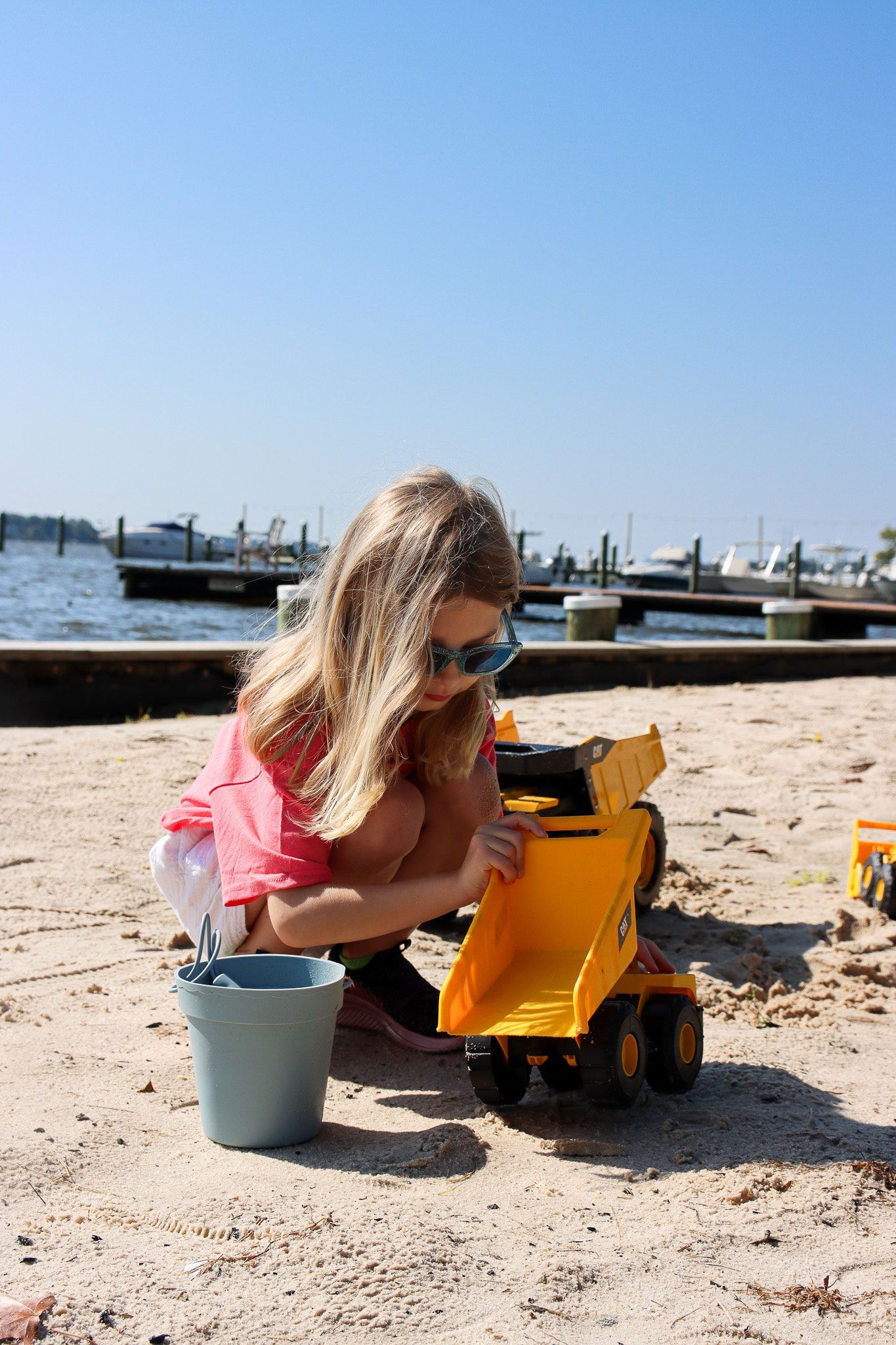 Child playing in sand beside a yellow toy dump truck at a beach, with boats and blue sky in the background