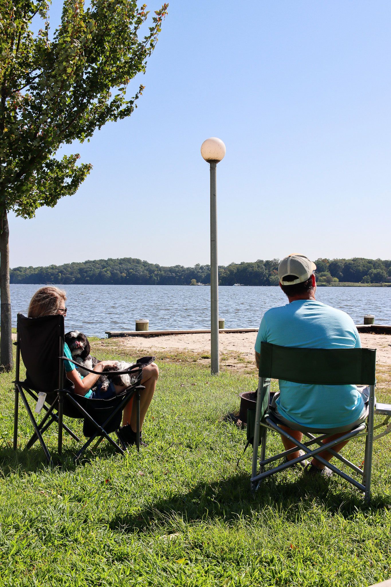 Two people sit in lawn chairs by a lakeshore under a clear blue sky.