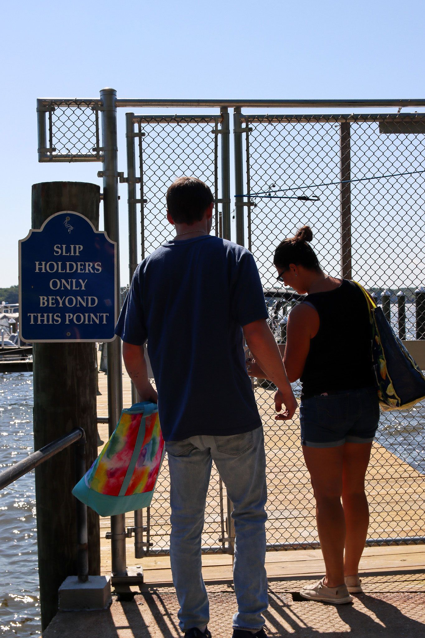 Two people stand at a chain-link gate on a sunny beach, one holding a colorful tote bag.