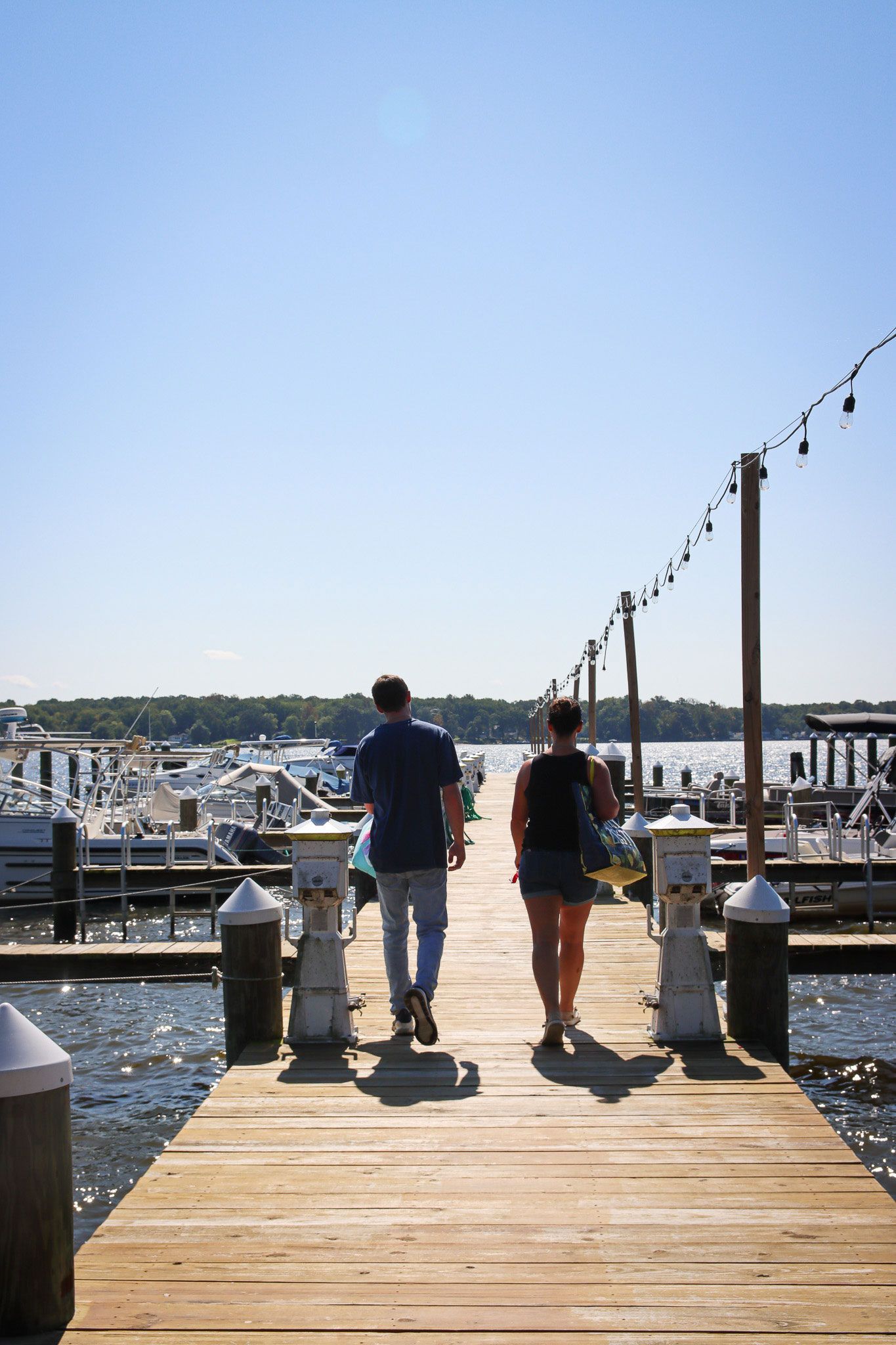 Two people walk down a wooden dock toward boats on a sunny marina