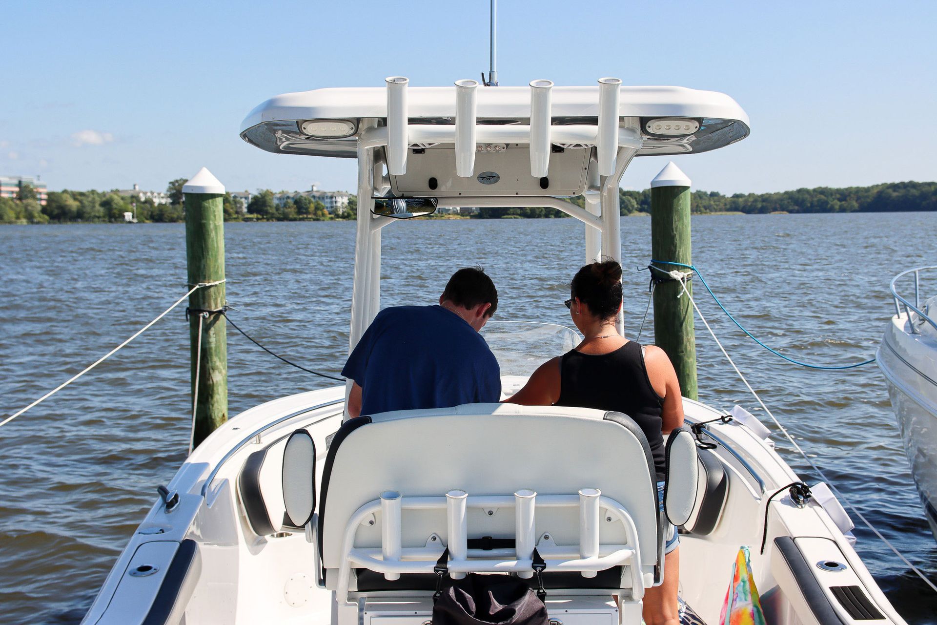 Two people sit on a boat deck facing the water under a white canopy.