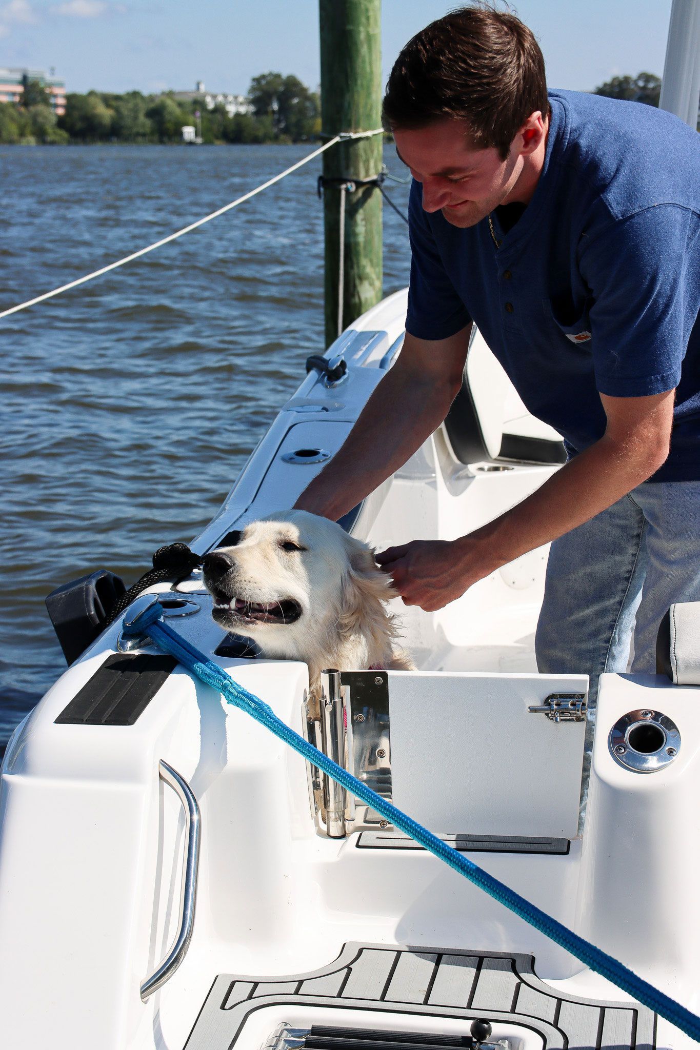Man on a boat leans over to pet a small dog beside a lake on a sunny day