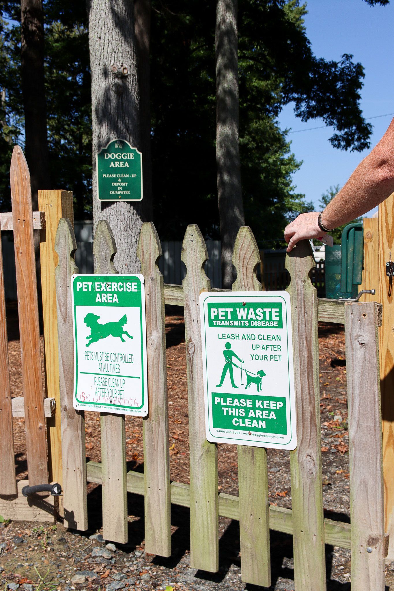 Green pet rules signs on a wooden gate, with a hand opening it near a fenced yard.